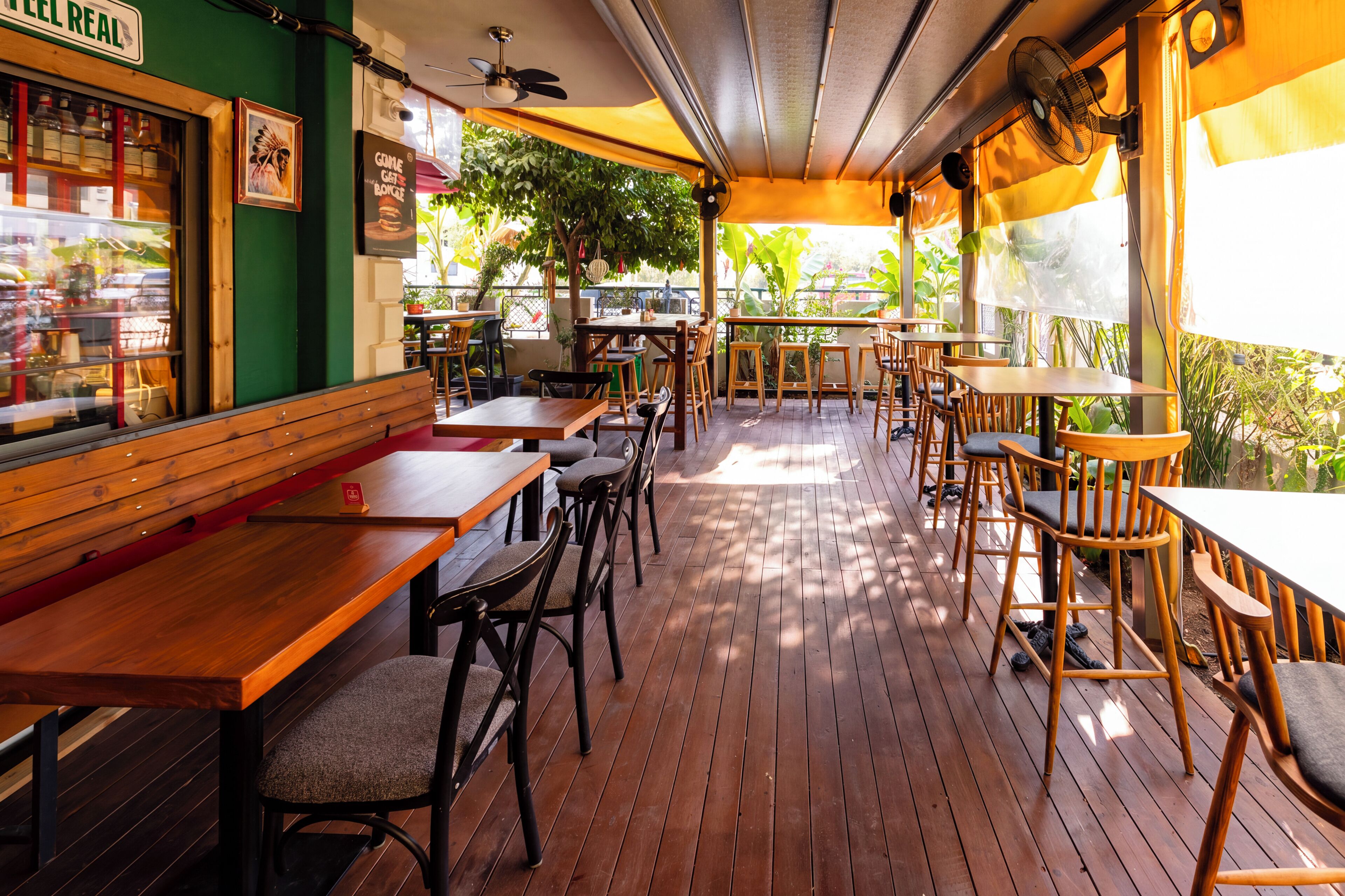 An outdoor cafe with wooden tables and chairs, a green wall, and a view of the street.