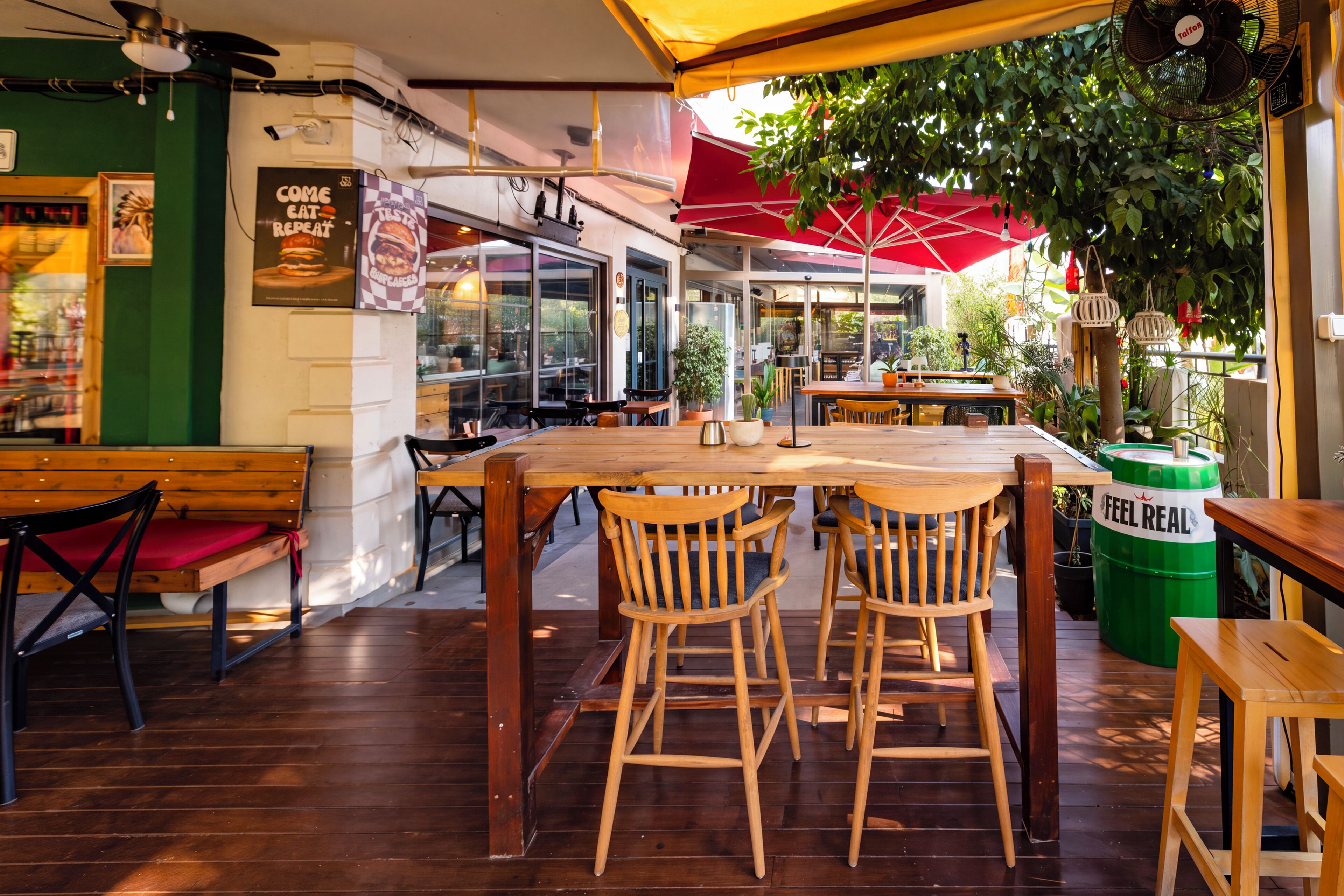 Un espace de restauration intérieur avec des tables et des chaises en bois, un parasol rouge et un tonneau vert portant l’inscription « FEL REAL ».