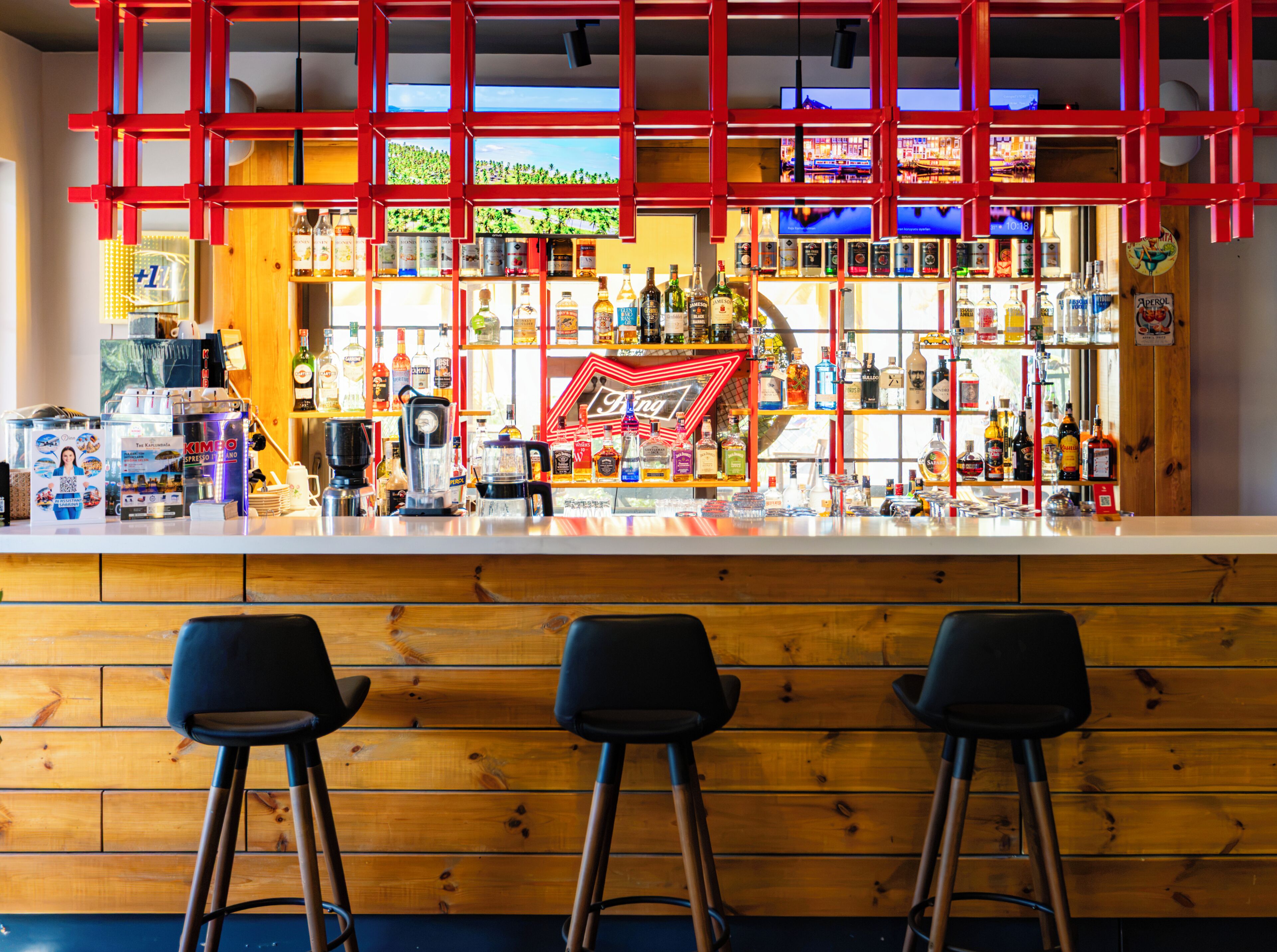 Uma área de bar com banquetas, um balcão com diversos tipos de bebidas e um letreiro de neon vermelho.