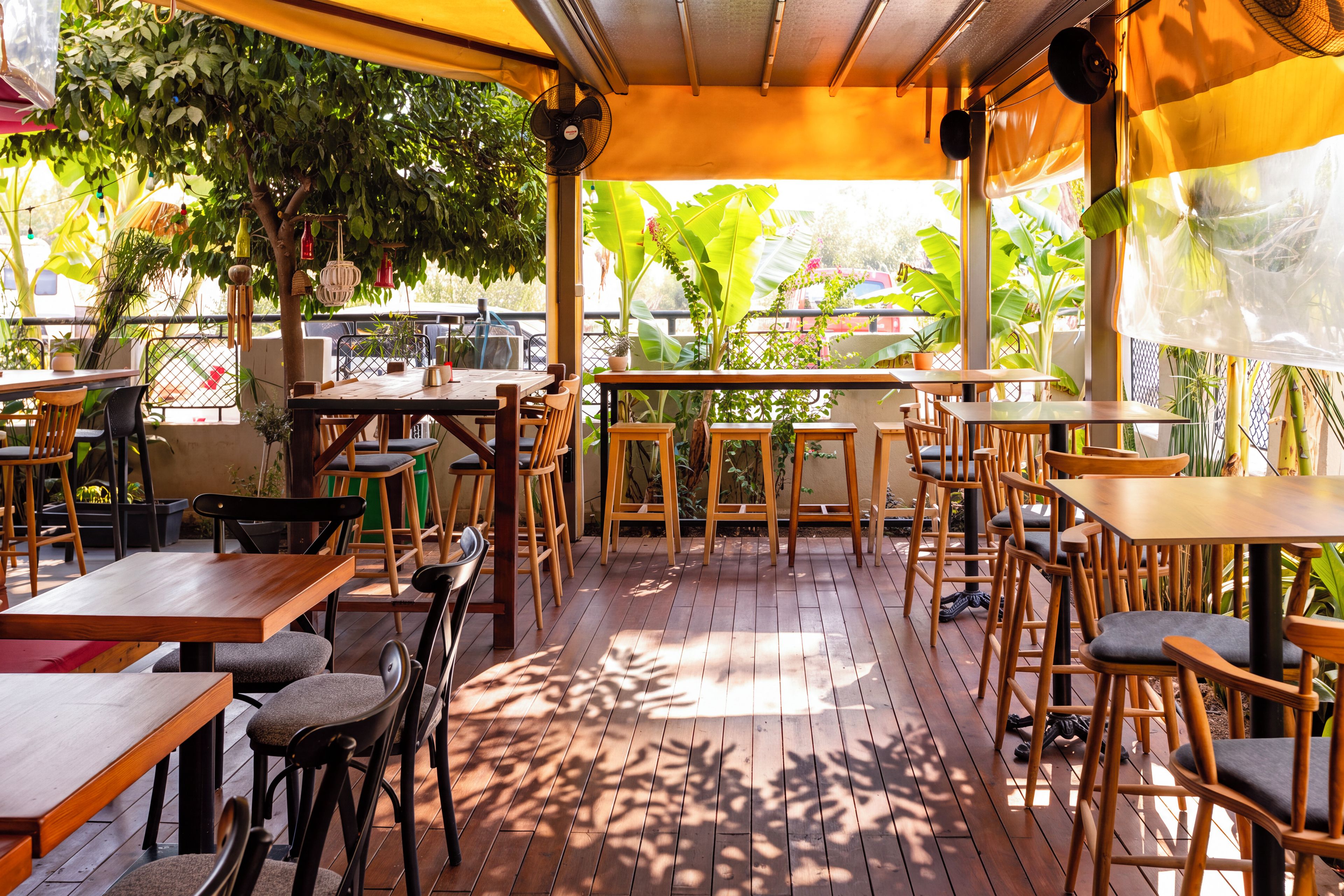 Un patio al aire libre con mesas y sillas de madera, rodeado de plantas en macetas y árboles.