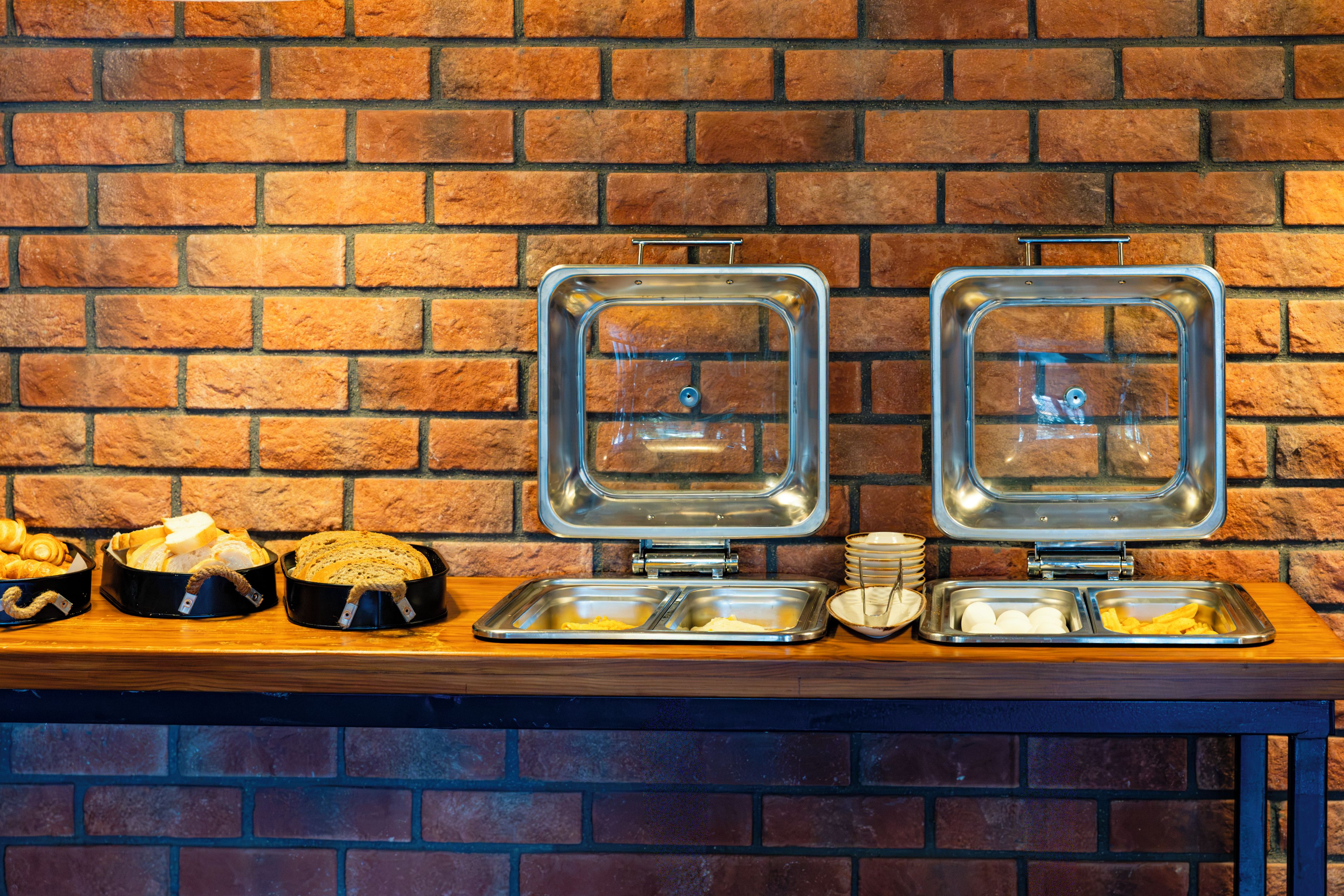 Een buffetopstelling met brood, eieren en andere ontbijtartikelen op een houten tafel tegen een bakstenen muur.