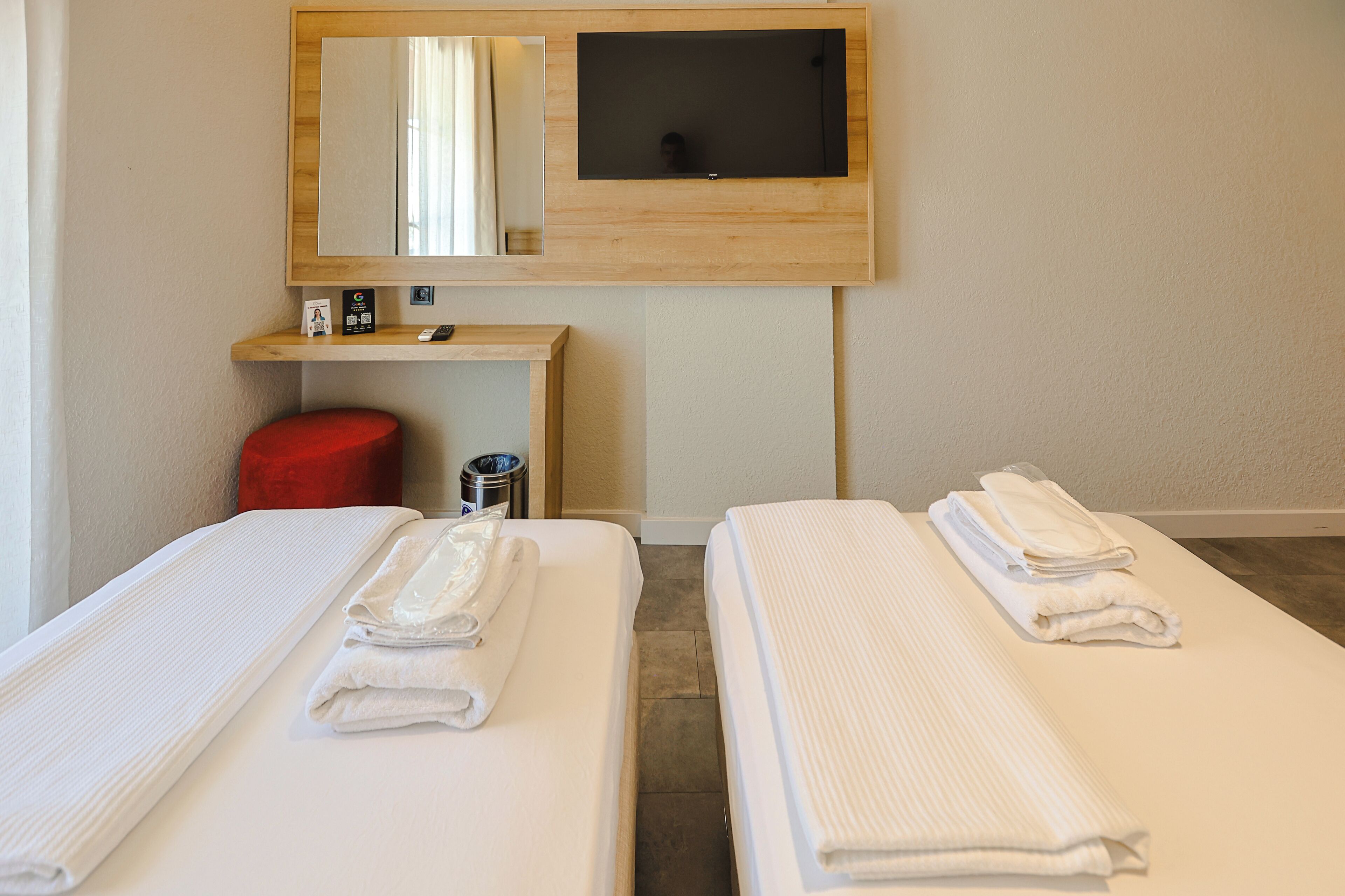 A hotel room with two beds, a wooden desk, a mirror, a TV, and a red stool.