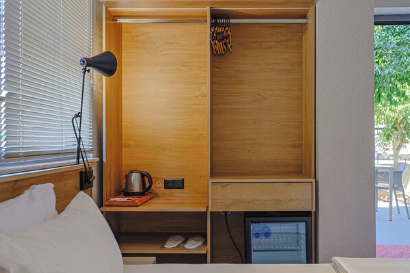 Une chambre de taille modeste, comprenant un lit, une petite table avec une bouilloire, un porte-chaussures et une fenêtre avec des stores.