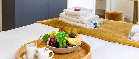 A modern hotel room with a kitchen area, a dining table with a tray of fruit and coffee cups, and neatly folded towels.