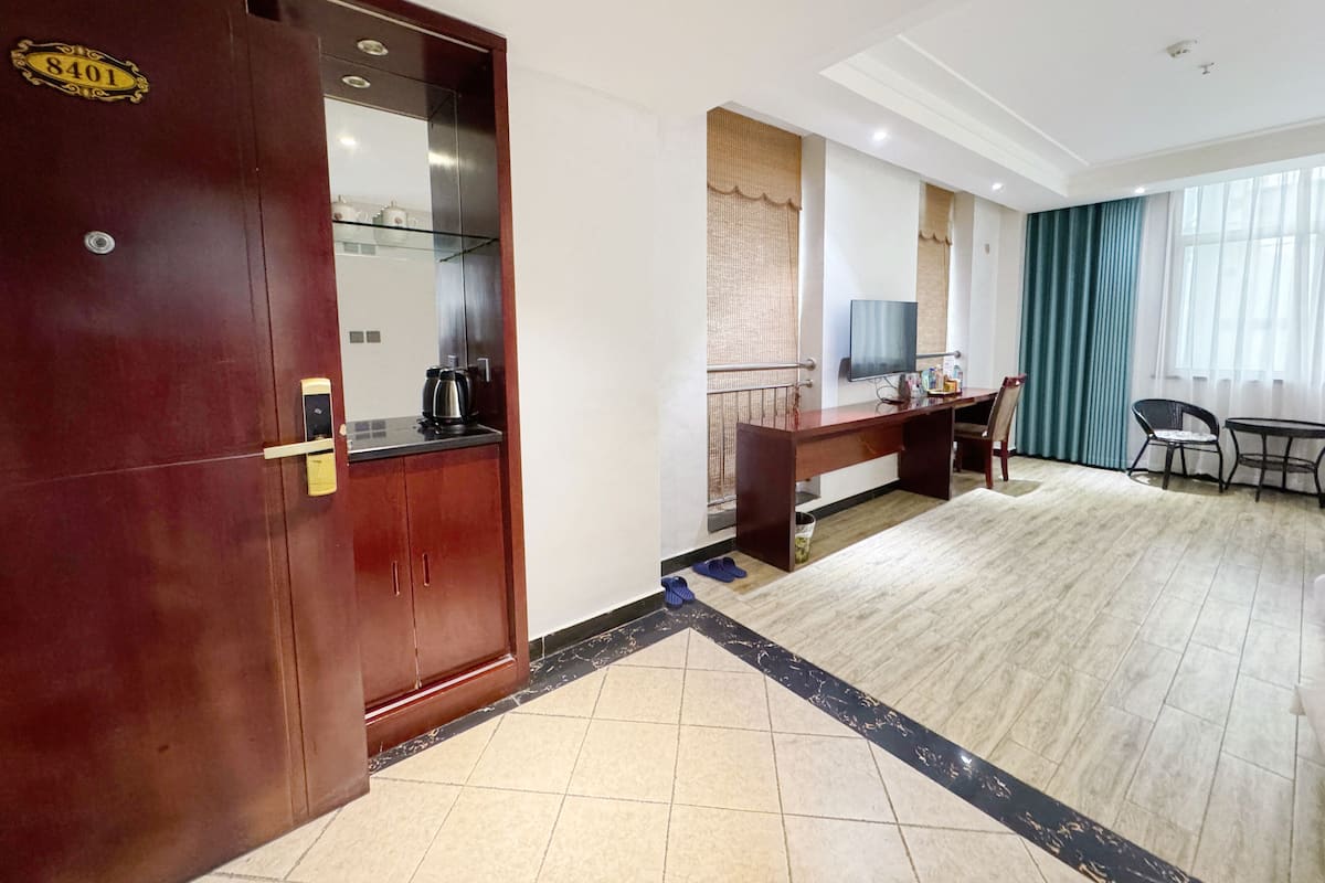 A hotel room with a wooden wardrobe, a desk with a television, a chair, and a large window with curtains.