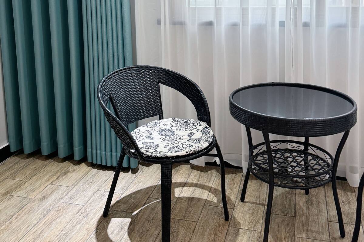 A room with a wicker chair and a small round table, a window with sheer curtains, and a teal-colored curtain.
