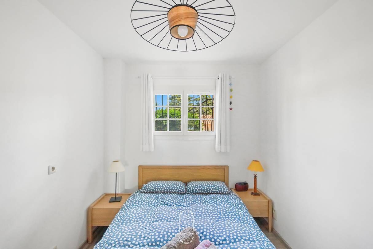 A bedroom with a bed, two bedside tables, a window with curtains, and a ceiling light fixture.