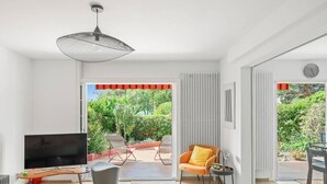 A modern living room with a TV, a sofa, a chair, and a dining table. There is a large window with a view of greenery outside. - Charmant RDJ T3 en bord de mer Carqueiranne (CARQUEIRANNE)