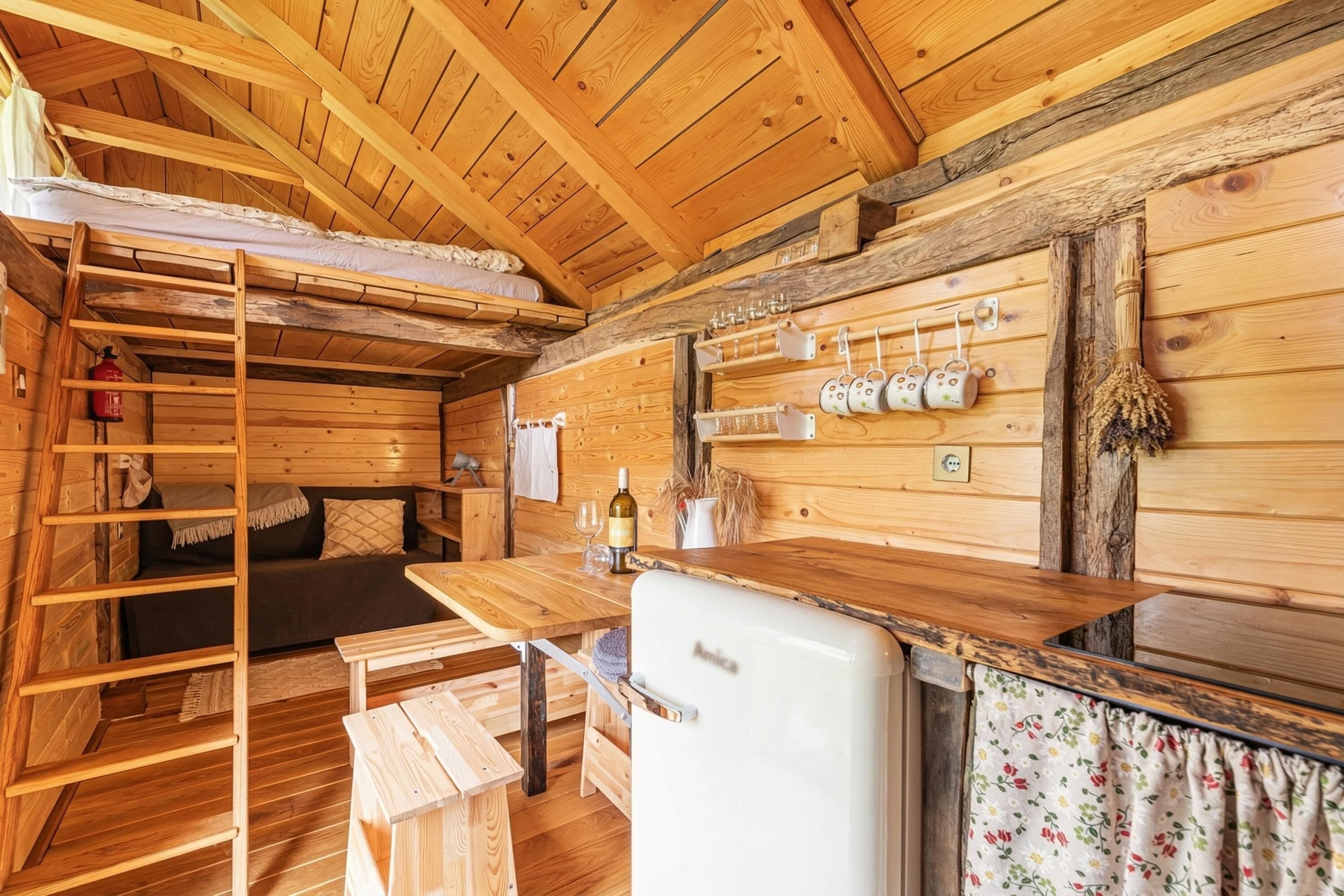 Interior de una cabaña de madera con literas, una zona de cocina con refrigerador y una escalera de madera.