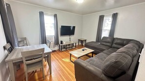 A living room with a sofa, a TV, a dining table, and chairs. - Strikeout - All Star Baseball Rentals (Oneonta)