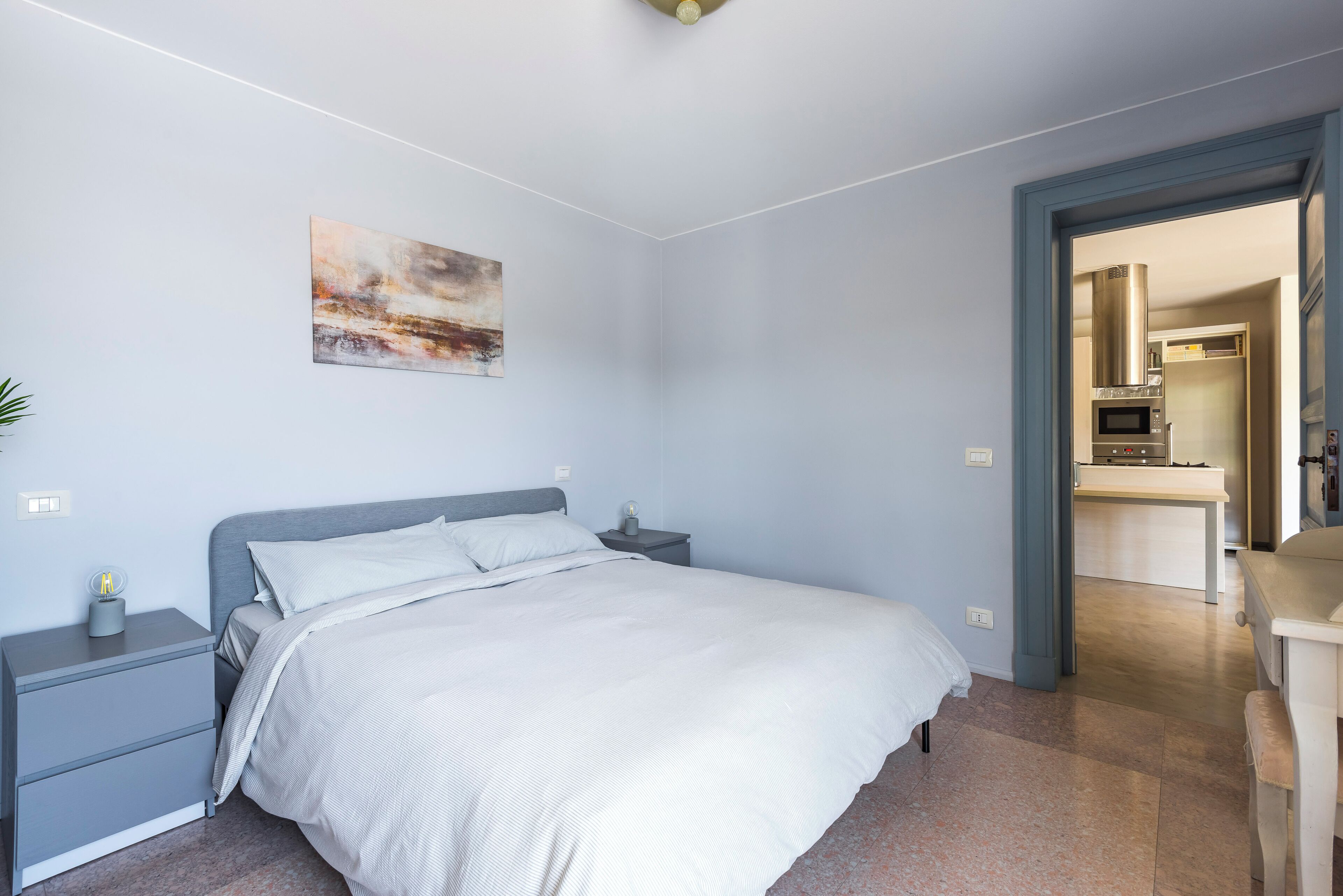 Un dormitorio con cama, mesitas de noche, un cuadro y vistas a la cocina.