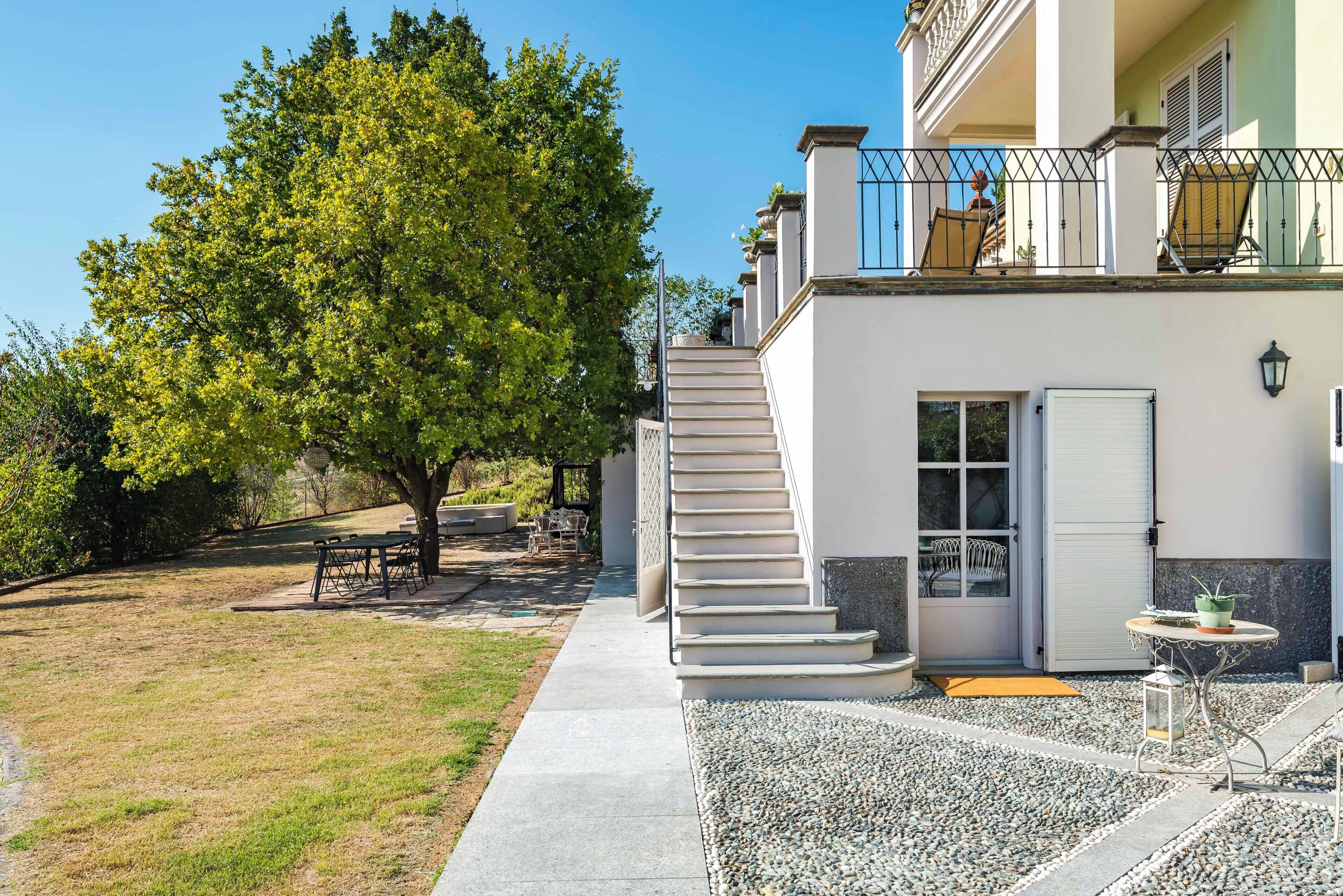 A two-story house with a staircase leading to a balcony, a small patio area with a table and chairs, and a tree in the yard.