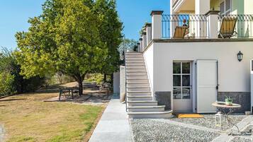 A two-story house with a staircase leading to a balcony, a small patio area with a table and chairs, and a tree in the yard.