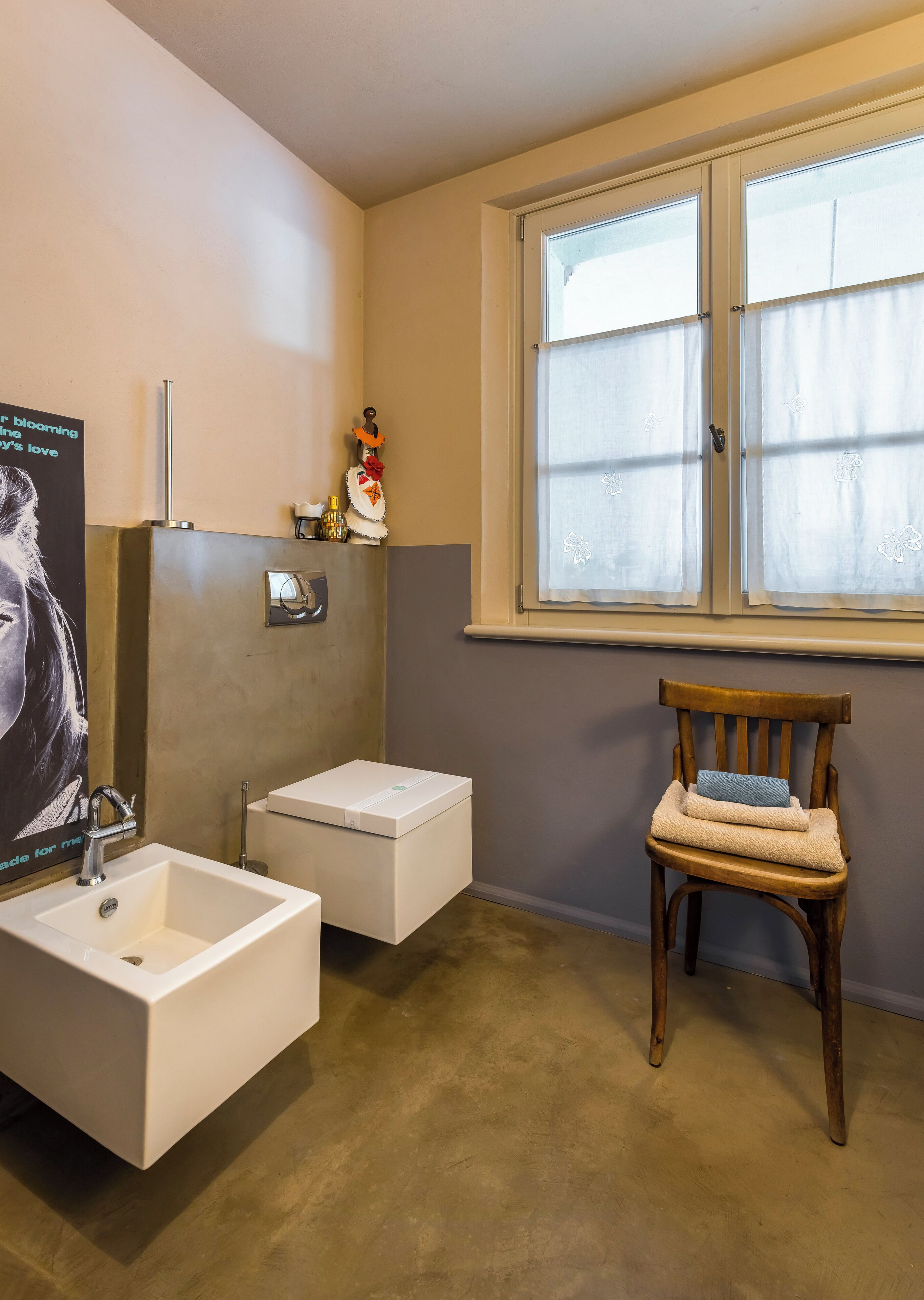 Une salle de bain avec un lavabo blanc, une chaise en bois et une fenêtre à carreaux dépolis.