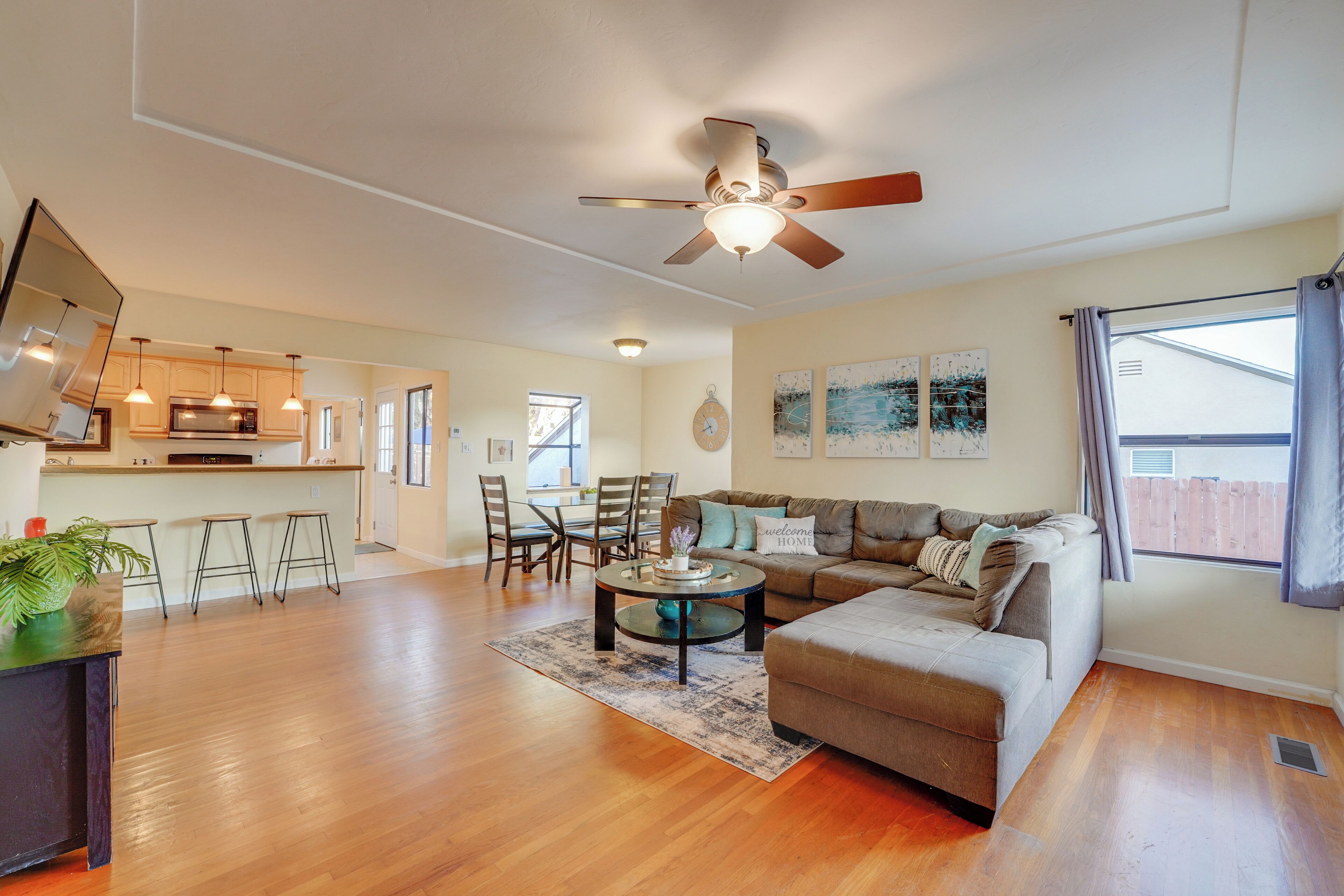 A spacious living area with a sofa, coffee table, and dining set. There is a ceiling fan, a kitchen area with cabinets and a microwave, and a window with curtains.