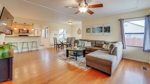 A spacious living area with a sofa, coffee table, and dining set. There is a ceiling fan, a kitchen area with cabinets and a microwave, and a window with curtains.