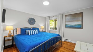 A bedroom with a bed, two bedside tables, a wall clock, a round wall decoration, a window with curtains, and a painting on the wall. - Short Description: Lakewood Bungalow | Screened Porch • 2 Baths • Basement Hangout • Walk to Lake• Onsite Laundry (Coloma charter Township)