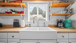 A kitchen with a white sink, wooden shelves, a window, and a coffee maker. - Short Description: Lakewood Bungalow | Screened Porch • 2 Baths • Basement Hangout • Walk to Lake• Onsite Laundry (Coloma charter Township)