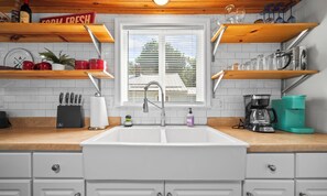 A kitchen with a white sink, wooden shelves, a window, and a coffee maker.