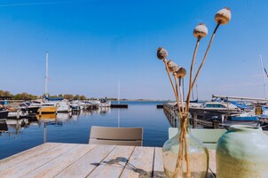 A wooden dock with a table and chairs, a vase with dried flowers, and a marina with boats in the background.