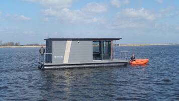 A stationary houseboat with a flat roof and a small deck, labeled "DE AMSTEL," on a calm body of water.