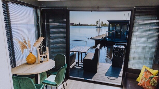 A modern interior with a dining area, a sofa, and a view of a water body with a dock. - Luxury Houseboat Getaway (Uitgeest)