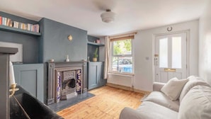 A living room with a fireplace, a sofa, a bookshelf, and a window with a view. - Cosy 2-Bed Cottage, close to Windsor By Tempstay (Berkshire)