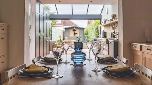 A dining table set for six with wine glasses and plates, a blue vase centerpiece, and a view of a garden through an open archway. - Cosy 2-Bed Cottage, close to Windsor By Tempstay (Berkshire)