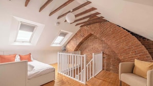 A room with a sloped ceiling, wooden beams, a brick wall, a white bed, a white chair, and a white railing. - Cosy 2-Bed Cottage, close to Windsor By Tempstay (Berkshire)