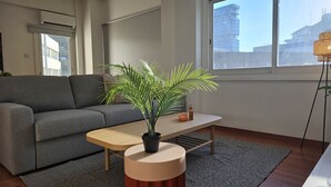 Een moderne woonkamer met een grijze bank, een houten salontafel, een plant in pot en een uitzicht op een stadsbeeld door grote ramen.