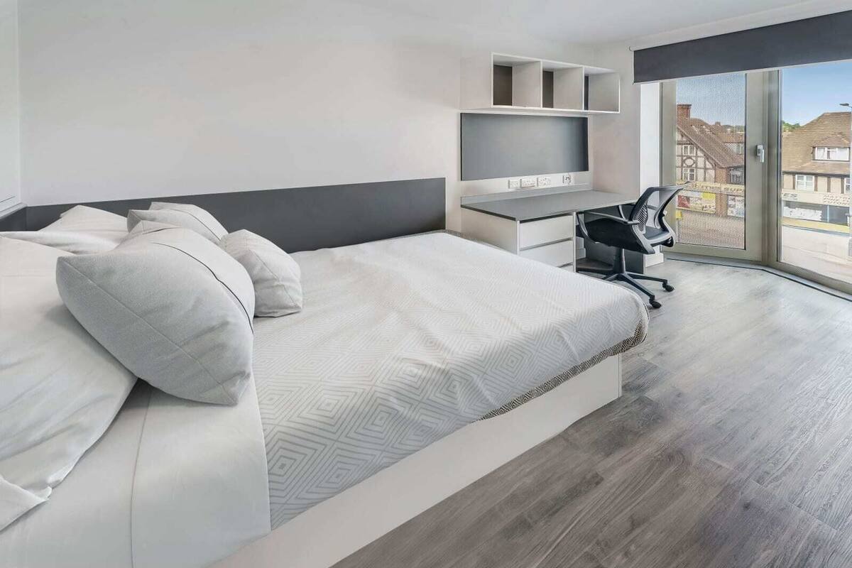 Ein modernes Hotelzimmer mit einem großen Bett, einem Schreibtisch mit Computer und Blick auf die Stadt durch große Fenster.