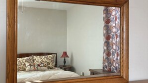 Ein Spiegel in Holzleiste auf einer Kommode, im Spiegelbild ein Bett zu sehen.
