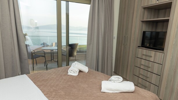 A hotel room with a bed, a TV, a balcony with a table and chairs, and a view of the sea. - SeaSide Village (Dirfys-Messapia)