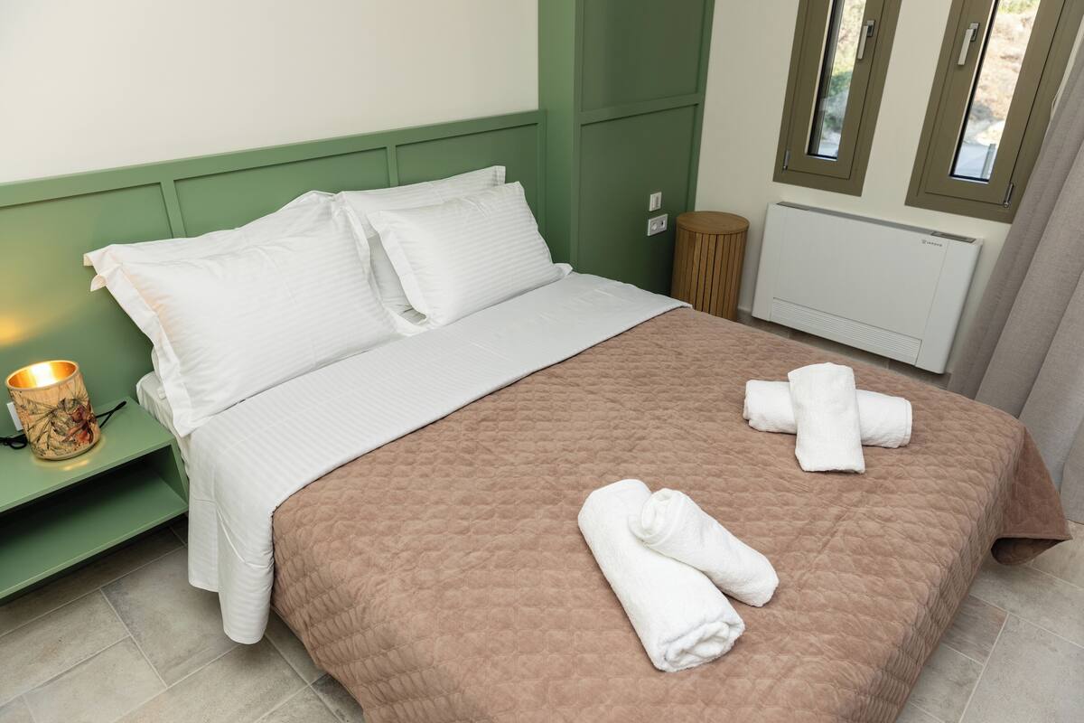Een opgemaakt bed met wit en bruin beddengoed, een bedlampje en opgerolde handdoeken op het bed.
