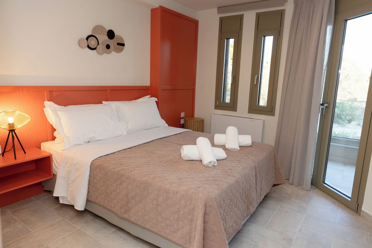 Een net opgemaakt bed met witte lakens en een beige sprei, een bedlamp en een decoratief object aan de muur.