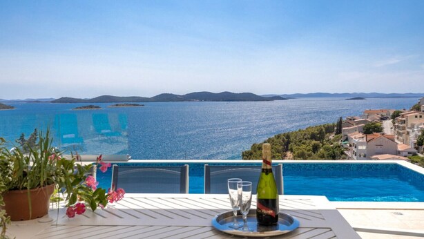 En terrass med en pool, en flaska vin och glas på ett brickbord, med utsikt över ett kustlandskap med kullar och en klar himmel.