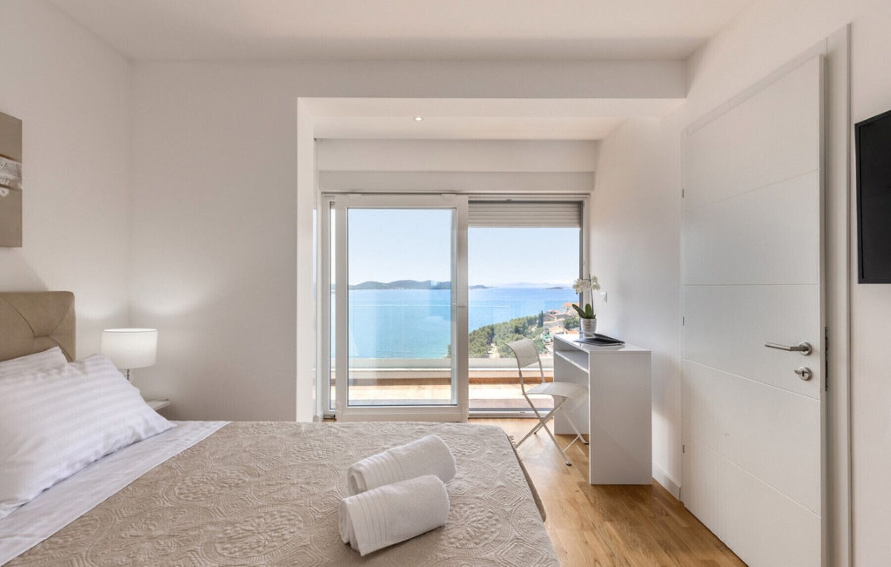 A bedroom with a large bed, a nightstand, a desk with a chair, and a view of the sea through a sliding door.