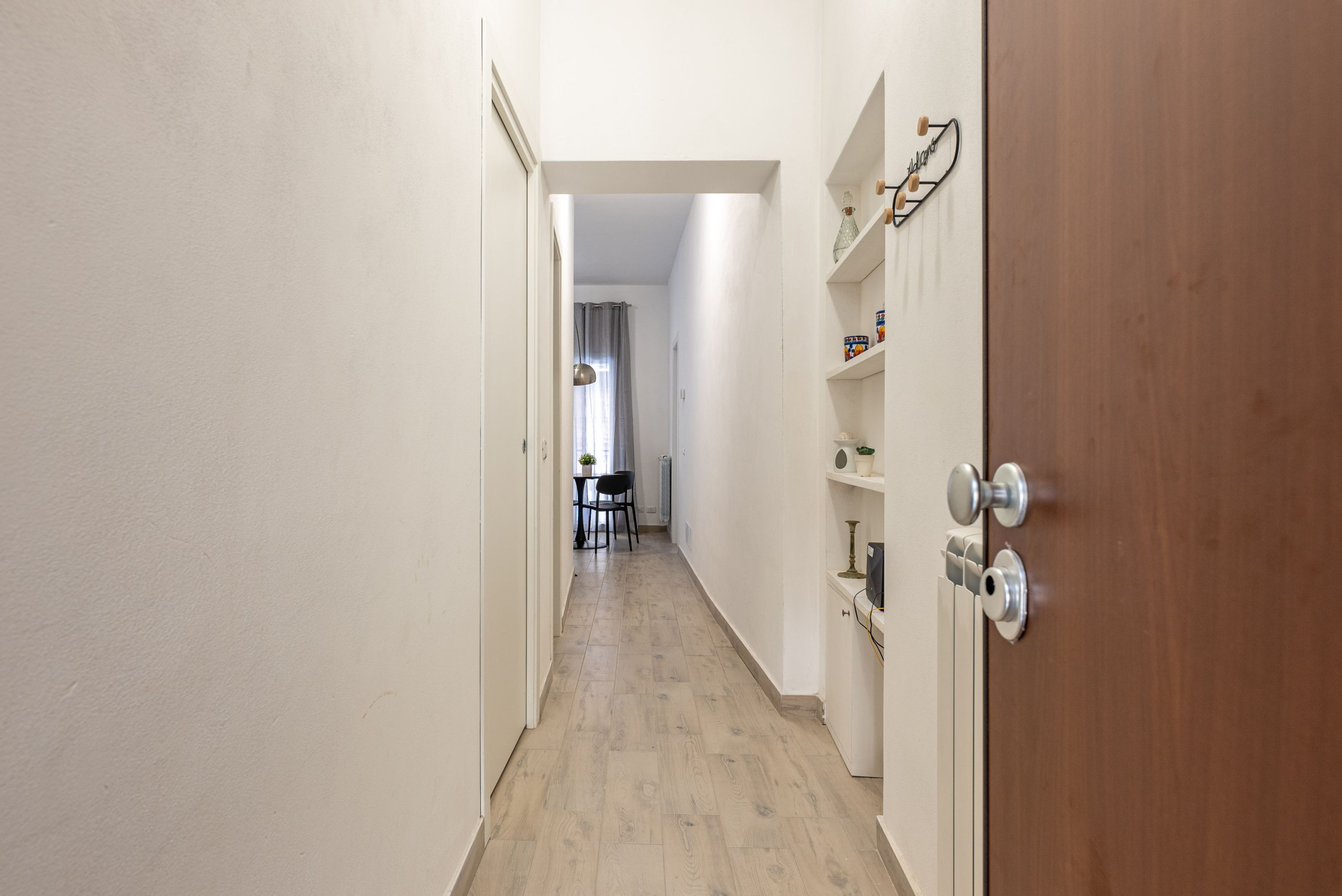 A narrow hallway with wooden flooring, white walls, and a wooden door with a metal handle. There is a small table with chairs in the distance.