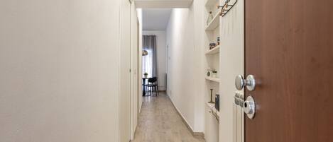 A narrow hallway with wooden flooring, white walls, and a wooden door with a metal handle. There is a small table with chairs in the distance.