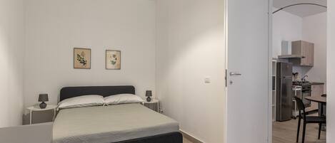 Ein Schlafzimmer mit einem Bett, Nachttischen und zwei an der Wand befestigten Bildern. Eine Tür führt zu einer Küchenzeile mit Kühlschrank und Herd.