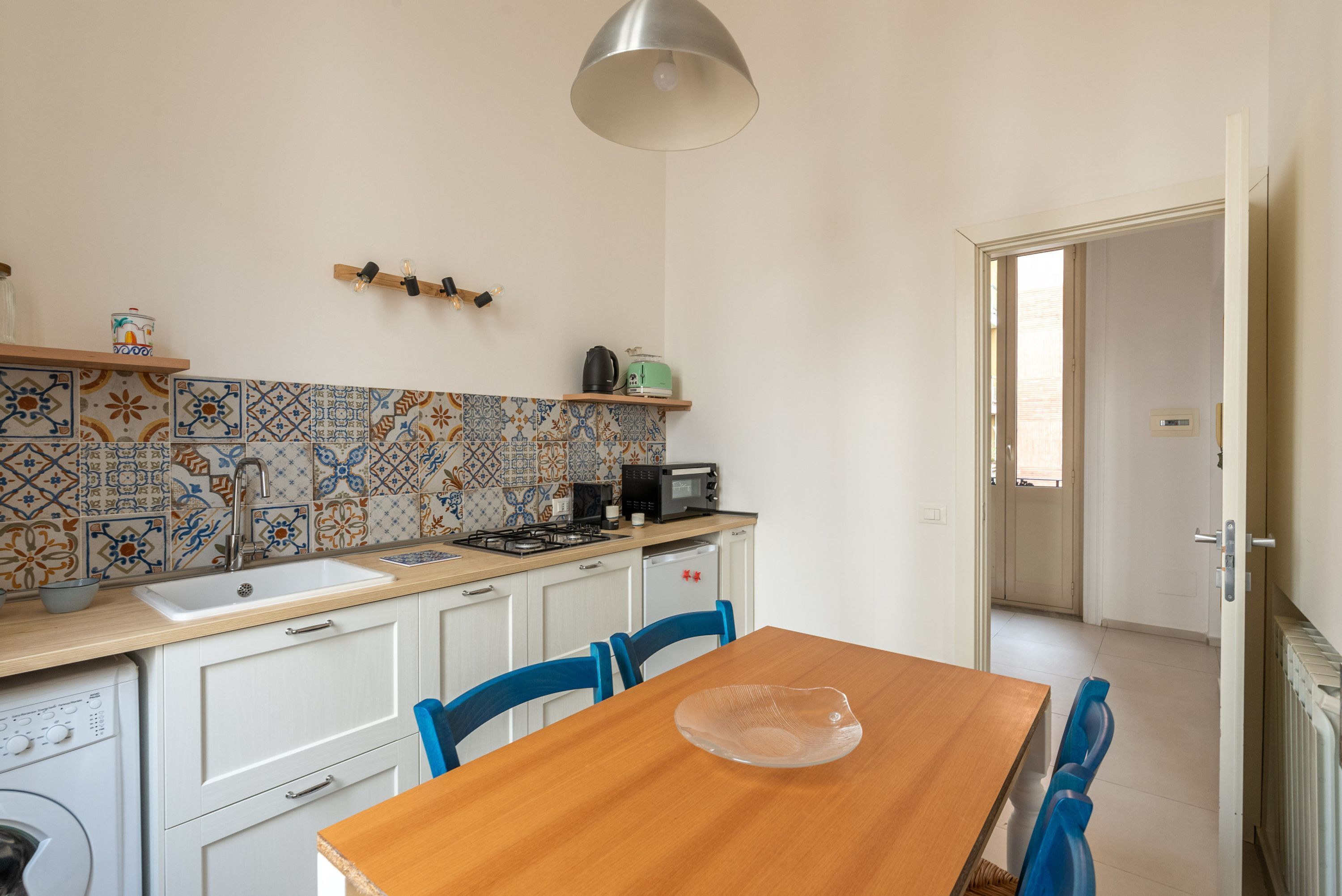 Uma cozinha compacta com uma mesa de madeira, cadeiras azuis, painel de azulejos e luminária pendente.