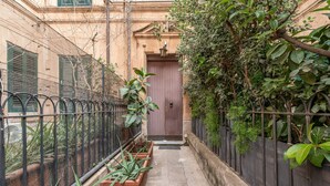 Une ruelle étroite, bordée d’une clôture en métal, avec des plantes en pot et une porte au fond.