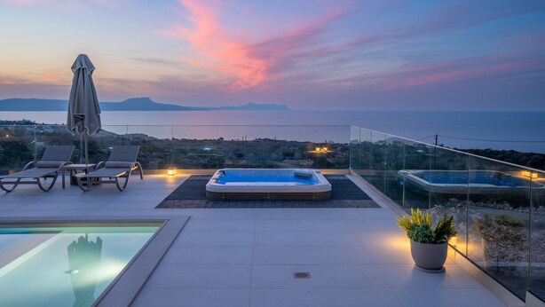 A rooftop pool area with a hot tub, lounge chairs, and a glass balustrade overlooking a body of water at sunset.