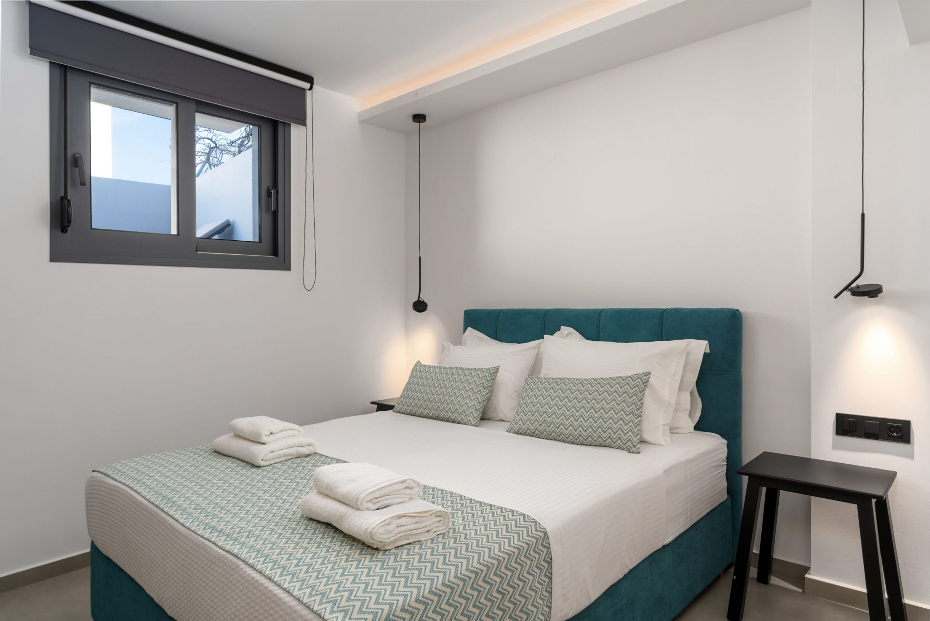 Quarto moderno com cabeceira verde-água, cama grande com roupa de cama estampada em branco e verde, mesa de cabeceira pequena e janela com vista para o exterior.