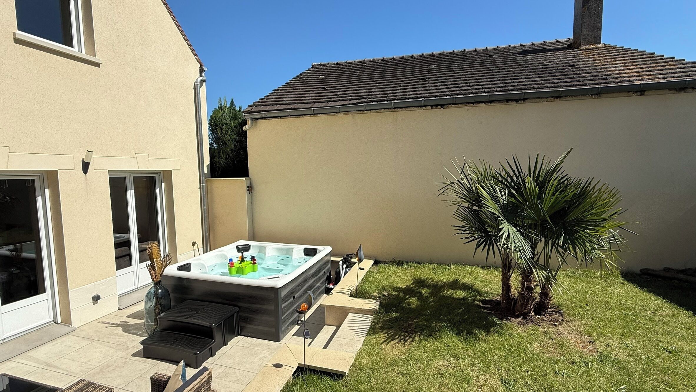 A backyard with a hot tub, a small palm tree, and a house with a tiled roof.