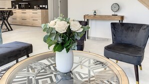 Un salon moderne avec une table ronde en verre, un vase blanc contenant des fleurs blanches, un fauteuil sombre et, au fond, une table à manger en bois.