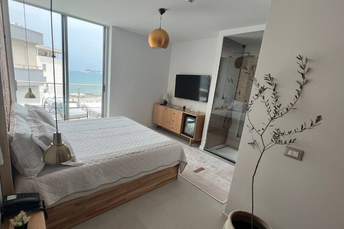 A bedroom with a large bed, a TV, a wooden dresser, and a view of the sea.