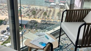 A view from a hotel room with a large window overlooking a beach. There is a wooden chair and a small table with a menu on it. - Luxury Rooftop Suites by Blumont (Rrashbull)