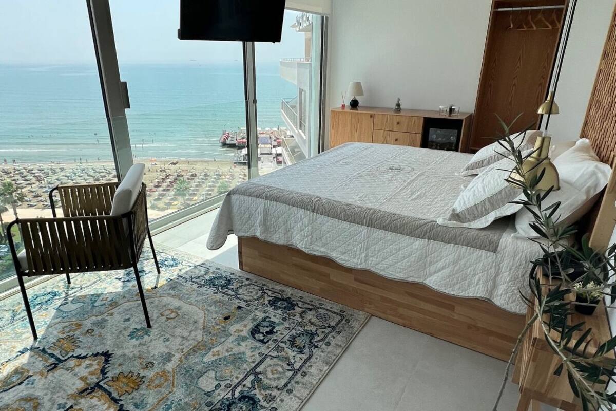 A bedroom with a large bed, a wooden headboard, a bedside table with a lamp, a TV mounted on the wall, and a view of the beach and ocean through large windows.