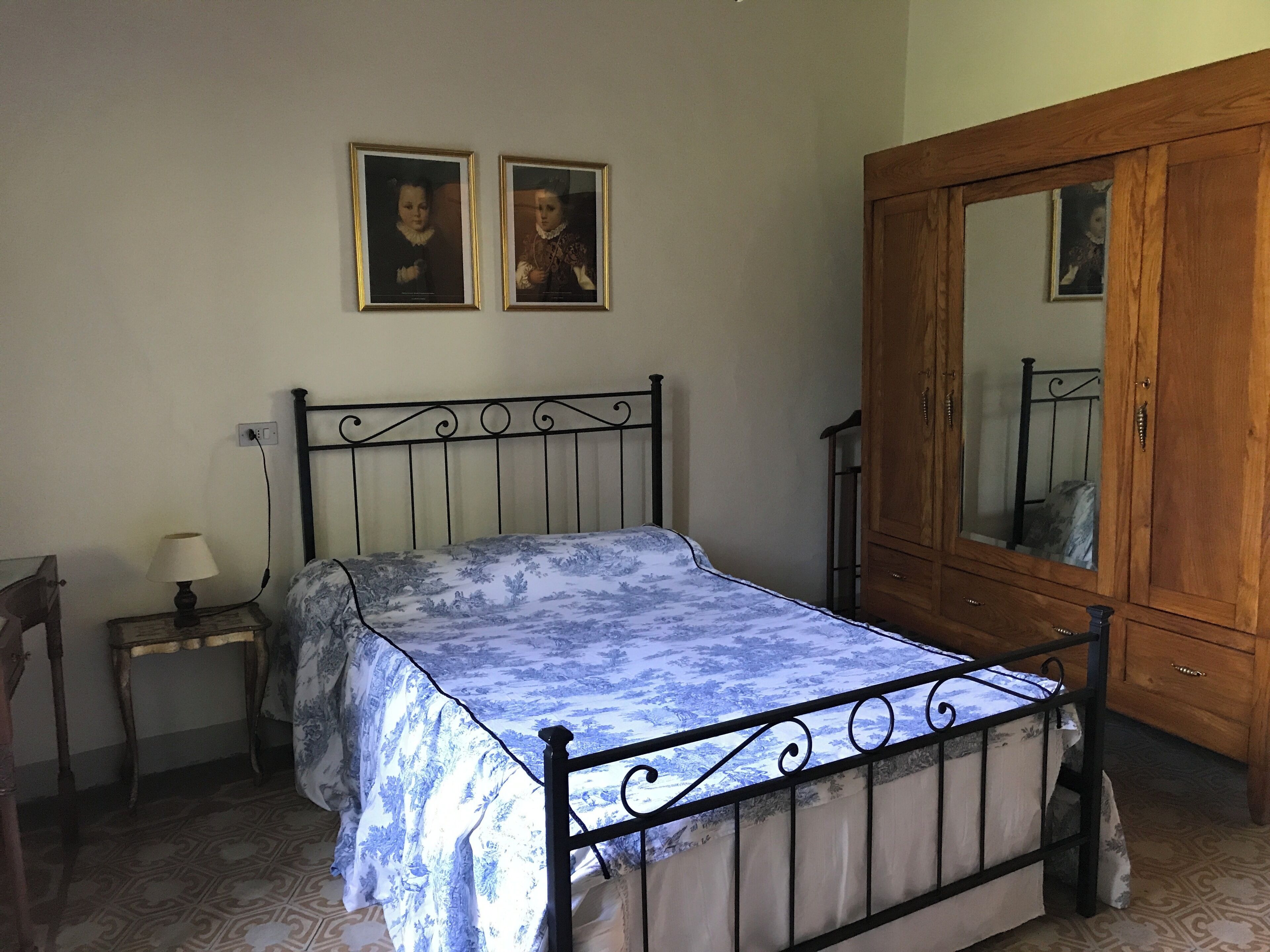 A bedroom with a bed, a nightstand, a lamp, a wooden wardrobe, and two framed pictures on the wall.